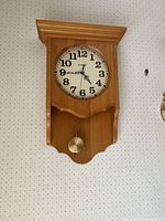 Antique wooden wall clock with white face, black Arabic numerals, black hands, and brass pendulum mounted on a white patterned wall.