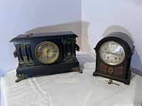Side view of both clocks on white cloth background showing ornate details and winding keys.
