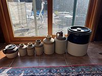 Six vintage ceramic crocks and jugs arranged side by side on a tiled floor near a window with outdoor view. The collection ranges from small to large sizes with different shapes and glaze patterns.
