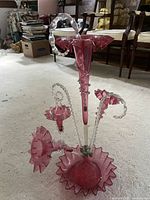 Front view of antique ruby glass epergne on carpet with furniture in background. Shows large central trumpet vase and three smaller side vases with clear twisted glass stems and ruffled edges.