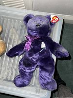 Purple Ty Beanie Baby teddy bear with a white embroidered rose on chest, purple satin ribbon bow, black button eyes, sewn black nose, and original Ty tag attached to ear.