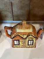 Front view of a ceramic tea pot shaped like a small cottage house with spout as chimney and handle shaped like a branch or wood, in brown and beige colors.