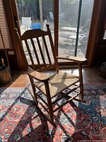 Photo of single vintage wooden rocking chair on patterned rug in front of glass door with outdoor view. Chair has vertical slats on backrest and woven fiber seat.
