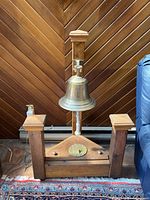Photo showing the full mounted bronze ship bell on its wooden frame against a wood-paneled wall with castors visible at the base and attached pull rope.