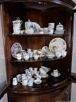 Wooden corner cabinet with multiple shelves holding an assortment of fine china porcelain teacups, saucers, teapots, creamers, sugar bowls, and decorative plates with floral and intricate designs.