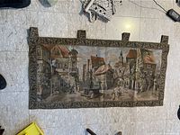 Photo showing the full antique tapestry laid flat on a tiled floor with visible hanging loops and detailed street scene design.