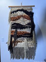 Front view of the hanging tapestry showing woven wool and yarn in abstract patterns with fringe at the bottom and wooden hanging rod.