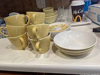 Stacked yellow porcelain coffee cups with handles, white bowls, and white plates with yellow floral decoration arranged on a table.