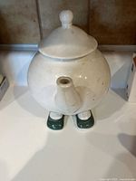 Front view of vintage off-white ceramic teapot with round body, lid with knob handle, short spout and two green shoe-shaped legs.
