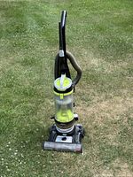 Front view of the upright Bissell Power Groom vacuum cleaner on grass, showing clear dust container and swivel head base.
