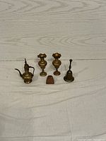 Five small brass items including a miniature pitcher, two matching candlesticks, a small handbell, and a small decorative brass piece displayed on a white cloth against a wooden backdrop.