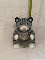 Gray and white plastic cat figurine holding a glass fish bowl, shown from a slightly angled front view with measuring tape behind for scale.