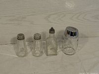 Four clear kitchen containers on white surface: two small shakers, one small bottle with tapered metal lid, one larger shaker with chrome metal lid.