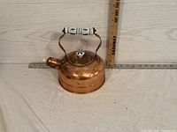 Side view of copper kettle showing ceramic handle and lid knob with blue floral design, placed against a wall with rulers for scale.