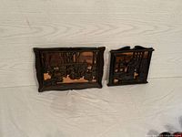 Photo of two copper backed wall art pieces side by side on a floor against a wall, showing their full front views with dark wooden frames and copper relief scenes