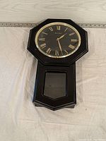 Full view of the black Daniel Dakota wall clock, showing the octagonal clock face with Roman numerals, gold hands, thick black frame, and lower pendulum glass window frame.