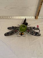 Front view of metal garden dragonfly with stained glass wings and green glass body with measurement scale showing approximately 13 inches wide.