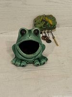 Close-up of the green ceramic frog sponge holder and frog-themed wind chime on a white surface against a light wood background.