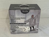 Box packaging showing model wearing Hoodzy Cozy Hooded Sweater Blanket with white and gray leopard print, describing product features: reversible with Sherpa inside, kangaroo pocket, one size fits all, unisex