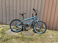 Blue BMX style bike leaning against a beige metal shed outdoors on grass and cement patch, showing full left side profile