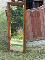 Large full-length mirror leaning against a wooden barrel outdoors, reflecting grass and trees. Medium brown thick wood frame visible.