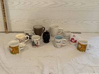 Wide shot showing all 11 assorted mugs arranged in front of a white wood paneled background and a wooden ruler for scale.