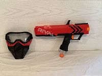 Red Nerf Rival Apollo Xbox-700 toy blaster and black and red protective face mask on light wood grain surface.