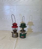 Photo showing two vintage Coleman lanterns side by side on a cream-colored cloth, one red and silver, the other green with gauge.