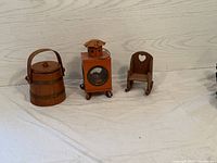 Three miniature rustic wooden items displayed on a white surface against a wood panel backdrop: barrel, lantern, rocking chair