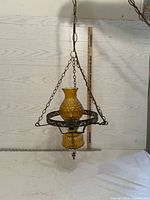 Full view of vintage amber glass hanging lantern with metal chains and decorative circular metal ring, against a wood-patterned backdrop with a ruler showing approximately 21 inch height.