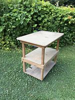 Wooden rolling cart with two lower shelves viewed at an angle outdoors on grass with greenery in background