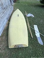 Yellow fiberglass boat hull upside down on grass along side wooden poles, sail canvas, rigging, and rudder.