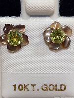 Pair of 10K gold flower-shaped earrings with green peridot stones in the center shown on a white holder with 10K gold stamp visible.