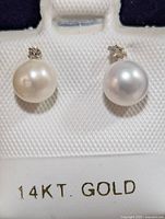 Front view of two 14K gold stud earrings each with a white freshwater pearl and a small diamond above it, set against a white textured card labeled 14KT GOLD.