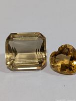 Photo showing both citrine gemstones side by side: rectangular emerald cut and heart-shaped cut, displaying color and facets.