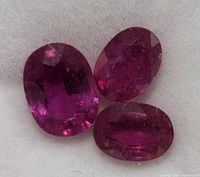 Close-up photo of three oval-shaped genuine enhanced rubies showing the deep reddish-pink color and faceted finish, placed on a light surface.