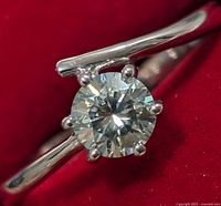 Close-up image showing the ring's split shank polished silver band and round moissanite stone held by six prongs on red velvet background.