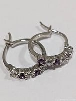 Close-up of one silver hoop earring showing alternating simulated alexandrite and clear stones, polished silver metal, hinged clasp closure.
