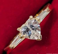 Close-up of silver ring showing heart-shaped clear cubic zirconia stone set in the center with tapered baguette CZ stones on each side against a red background.