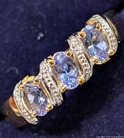 Close-up of the top of the silver ring featuring three oval light bluish-purple tanzanite stones with small clear accent stones on curved settings.