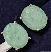 Pair of oval dyed emerald earrings with silver prong settings shown close-up on dark blue background.