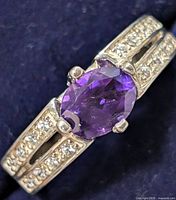 Close-up of sterling silver ring with central purple amethyst-colored CZ and clear cubic zirconia accents on band, displayed on dark fabric background.