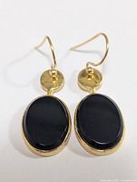 Front view of one earring showing the black oval onyx stone set vertically in a gold-tone silver bezel, with a round hammered gold-tone disk above and curved hook attachment.