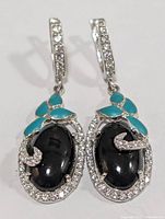 Front view of silver earrings with large oval black onyx stones, surrounded by clear cubic zirconia and turquoise inlay on top. Hinged clasp also decorated with clear CZ stones.