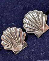 Close-up image of pair of silver earrings shaped like seashells with ridged design on a dark fabric background.
