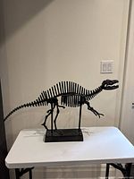 Full side view of a black metal dinosaur skeleton sculpture sitting on a white table against a light wall.
