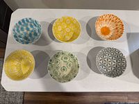 Top view of six stoneware bowls arranged in two rows of three showing colorful interiors with different patterns: blue checkered, yellow floral, orange radial, yellow striped, green floral, and gray geometric.