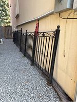 Wrought iron fence panels aligned beside a building wall on a gravel ground, showing traditional scroll and picket black iron design, with round finial post tops.