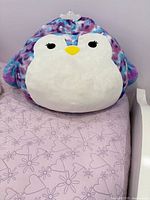 Front view of the large penguin Squishmallow on a lavender bedspread with heart and floral patterns. The penguin has a white face and belly, purple and teal mottled body, yellow beak, and black eyes with eyelashes. A silver crown accessory is visible on top of its head.