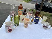 Front angle view of 16 assorted vintage glassware including firefighter and brewery mugs, steins, and bottles arranged on white tablecloth outdoors.